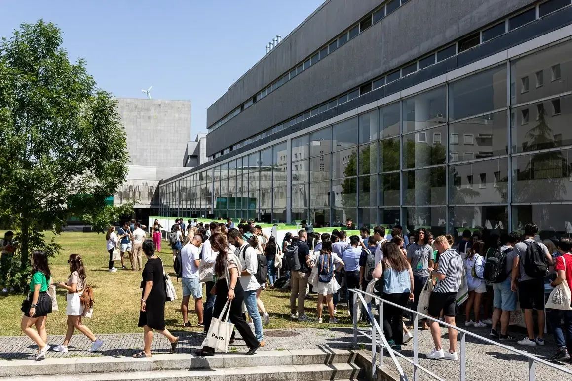 A imagem mostra um grande grupo de jovens reunidos no exterior de um edifício moderno, provavelmente num campus universitário ou num espaço institucional