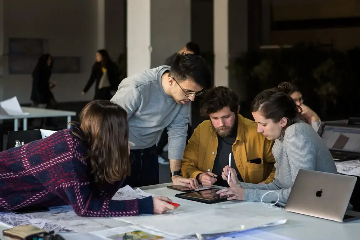 A imagem mostra um grupo de quatro pessoas colaborando intensamente numa mesa de trabalho. Elas parecem estar envolvidas num projeto criativo ou técnico