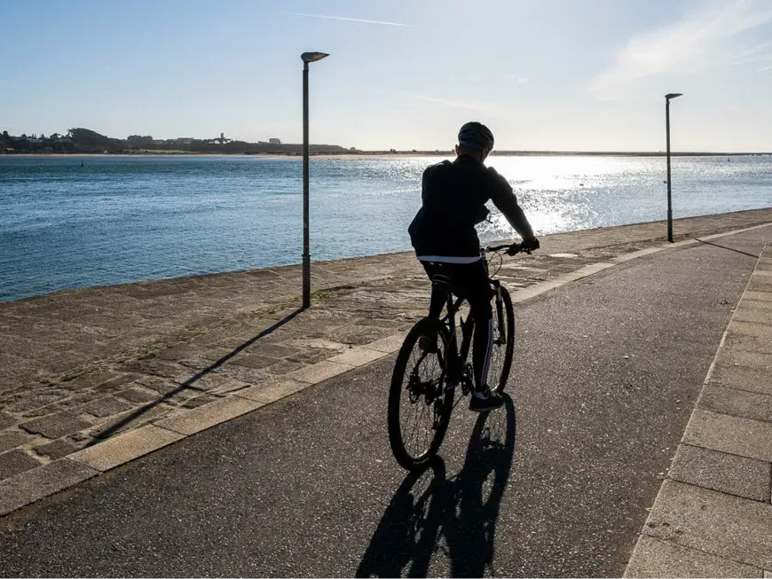 A imagem mostra uma pessoa a andar de bicicleta junto a uma zona costeira, provavelmente num passeio marítimo