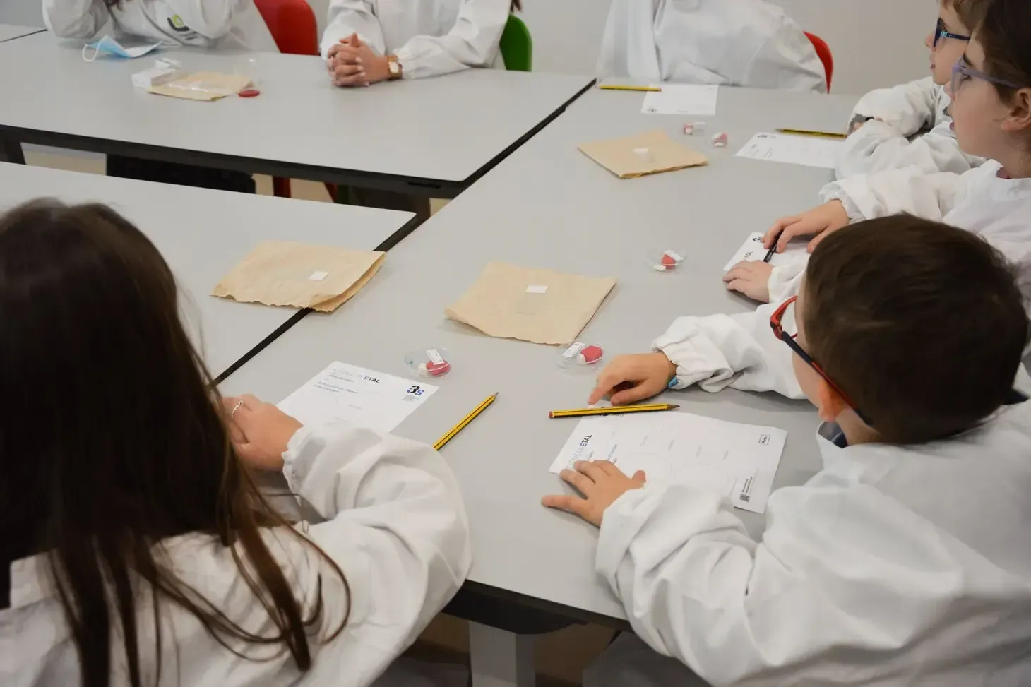 A imagem mostra um grupo de crianças sentadas ao redor de mesas retangulares, formando um grande conjunto de trabalho