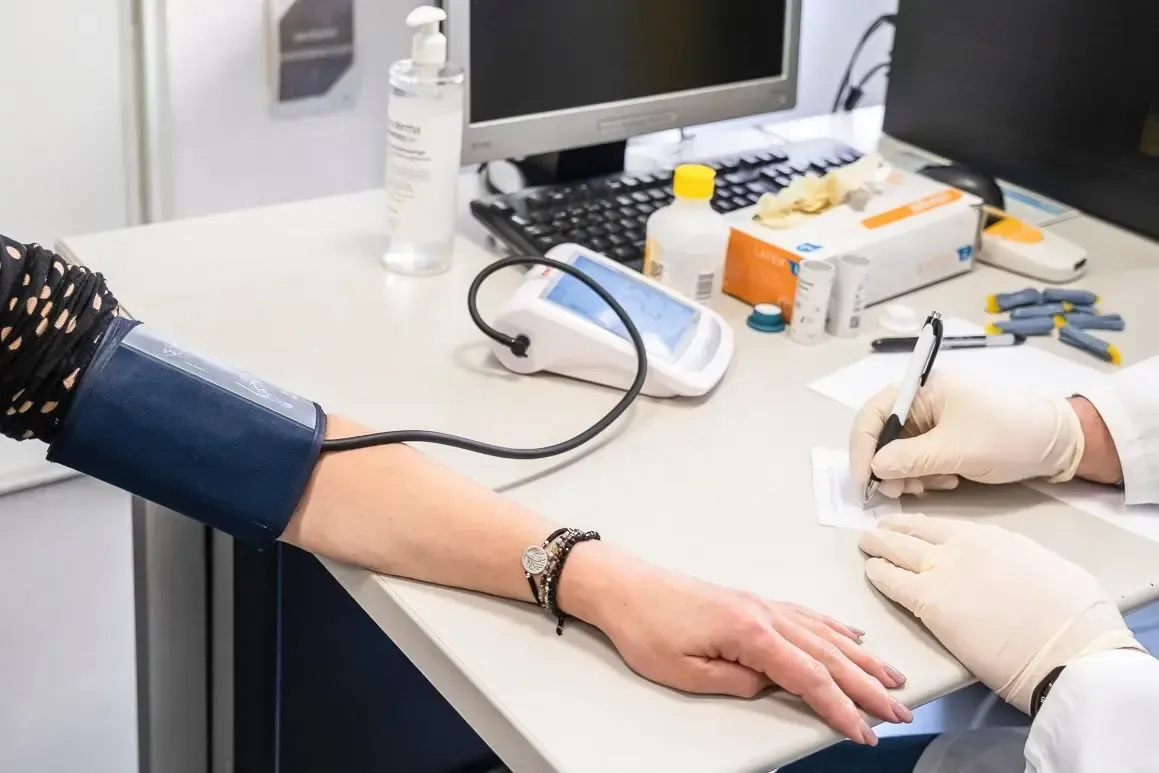 Na imagem, vê-se uma consulta médica ou um procedimento de rotina. O foco está no braço de uma pessoa que está a ser medida a pressão arterial com um braçadeira azul ligada a um tensiómetro eletrónico sobre a mesa