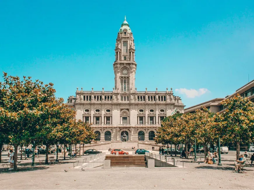 A imagem mostra uma praça ampla e luminosa, com céu azul claro e tempo ensolarado. Ao fundo destaca-se um edifício imponente de arquitetura clássica, a Câmara Municipal do Porto