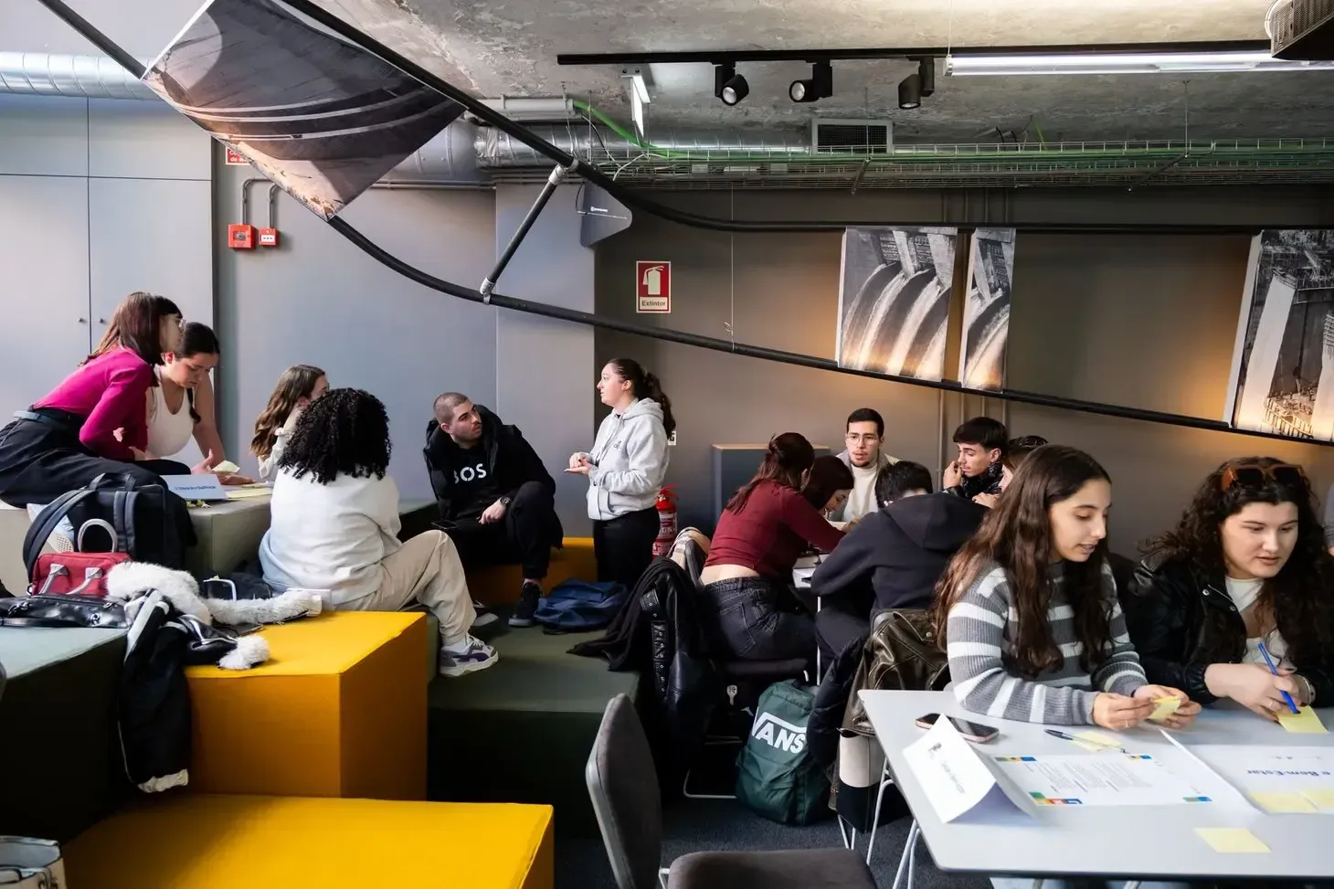 A imagem mostra um grupo de jovens reunidos num espaço interior moderno, com diferentes pontos de trabalho distribuídos pelo ambiente