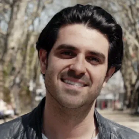 A foto mostra um homem jovem sorridente, ao ar livre, numa rua ladeada por árvores 