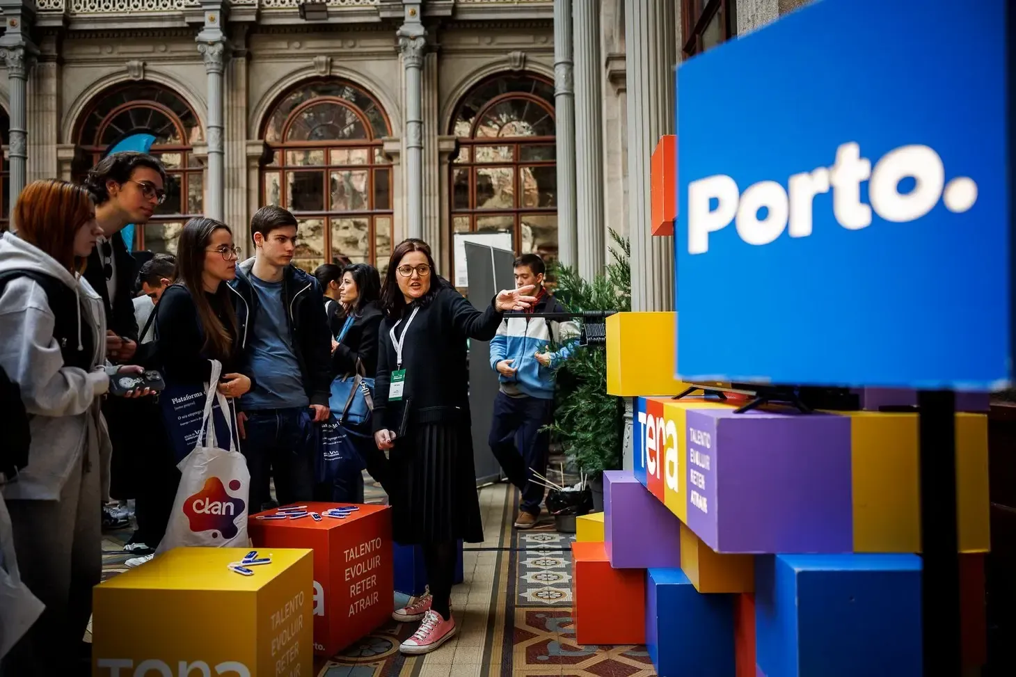 Na imagem vê-se um ambiente de evento ou feira num espaço interior histórico, com colunas e janelas altas ao fundo