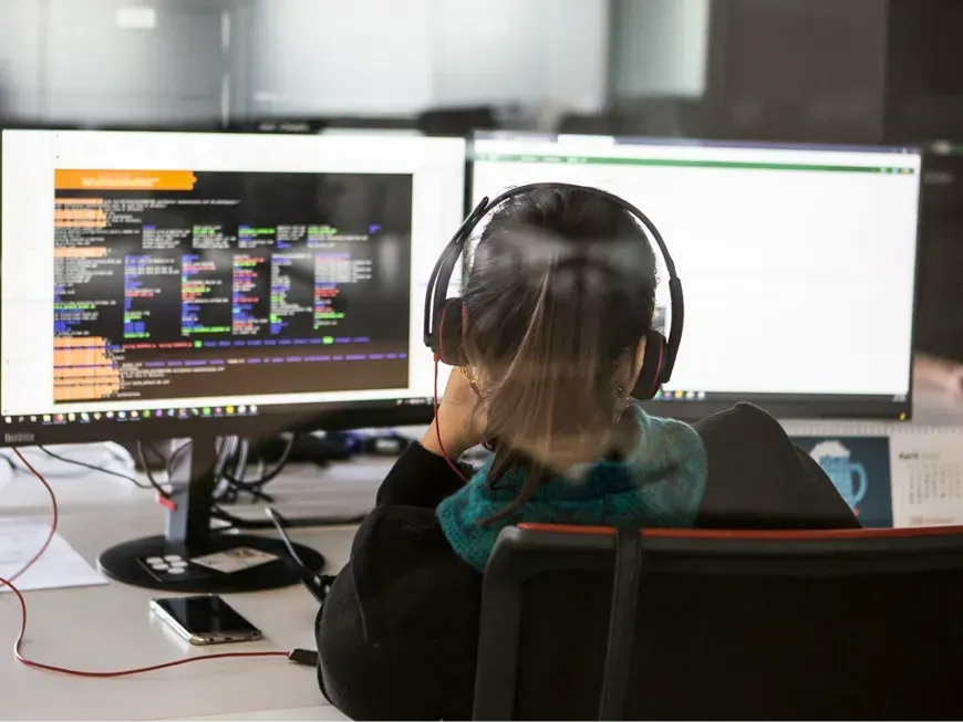 A imagem mostra uma pessoa sentada numa secretária, vista de trás, a trabalhar em dois monitores