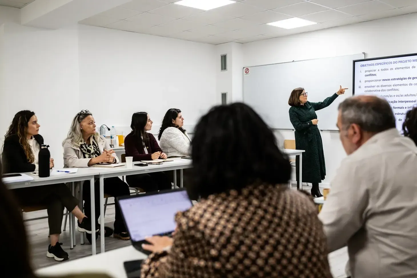 A imagem mostra uma sala de aula ou sala de formação com várias pessoas sentadas em mesas dispostas em formato de “U”