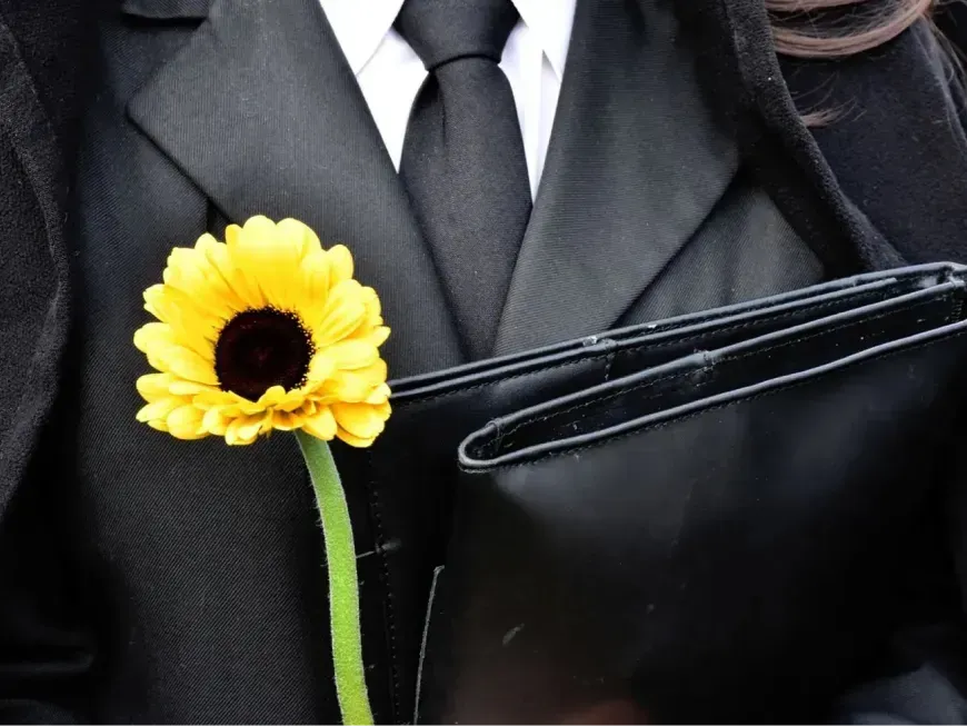 A imagem mostra uma pessoa vestida com traje formal preto — casaco, camisa branca e gravata preta — segurando uma pasta ou capa igualmente preta