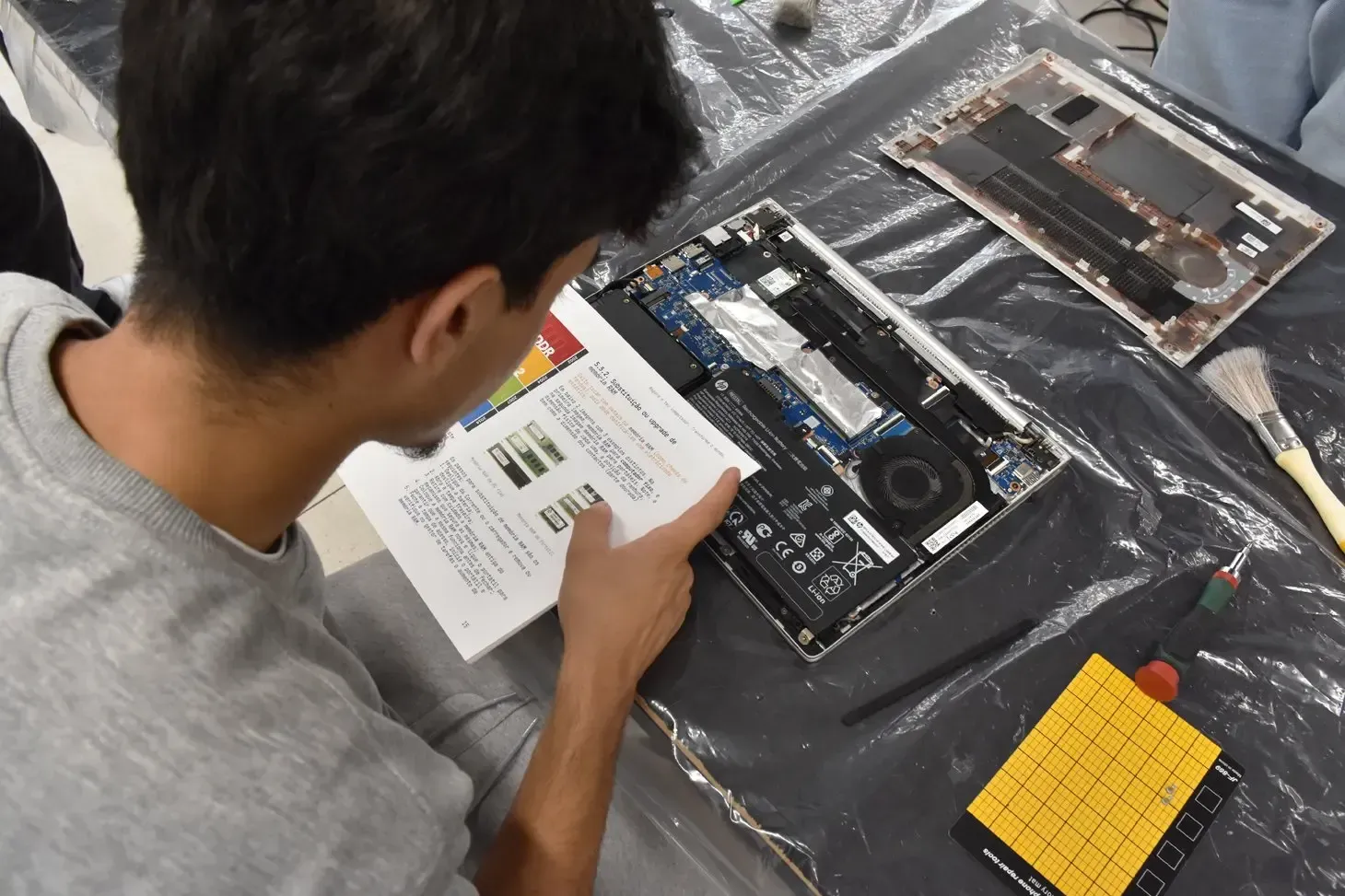 A imagem mostra um jovem sentado a trabalhar na reparação de um portátil. Ele observa atentamente um manual ou guia técnico enquanto aponta para uma das instruções, sugerindo que está a seguir um passo-a-passo